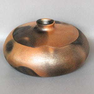 May include: A handcrafted ceramic container with a lid. The vessel is a warm copper color with dark brown speckled accents. The lid has a small, cylindrical handle. The overall shape is round and low, with a smooth, metallic finish.