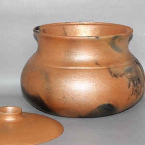 May include: A handcrafted, round ceramic jar with a lid. The jar is a warm, metallic copper color with dark, mottled accents. The lid is flat with a small, raised handle. The jar's shape is rounded with a slightly flared rim.