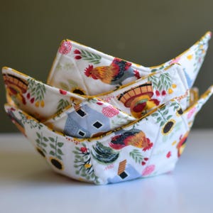 May include: A stack of four fabric bowl cozies. The cozies are white with a colorful rooster, sunflower, and farmhouse print. The edges are trimmed with yellow fabric. The cozies are designed to hold bowls of hot food.