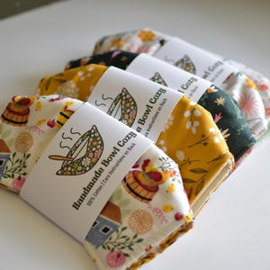 Peut inclure: Plusieurs coussins de bol faits &agrave; la main avec des motifs floraux. Chaque coussin est envelopp&eacute; d'une &eacute;tiquette blanche indiquant "Handmade Bowl Cozy" et comprend des instructions d'entretien. Les motifs incluent une maison, des fleurs et d'autres motifs.