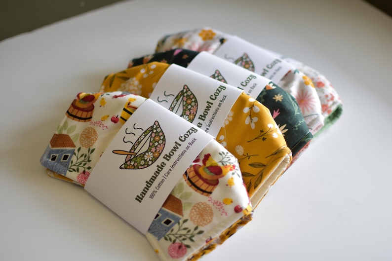 Peut inclure: Plusieurs coussins de bol faits &agrave; la main avec des motifs floraux. Chaque coussin est envelopp&eacute; d'une &eacute;tiquette blanche indiquant "Handmade Bowl Cozy" et comprend des instructions d'entretien. Les motifs incluent une maison, des fleurs et d'autres motifs.