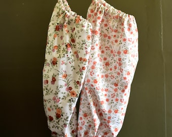 May include: Two fabric produce bags with floral patterns. The bags are white with a repeating pattern of orange, pink, and burgundy flowers and green leaves. The bags are hanging from a wooden rack.