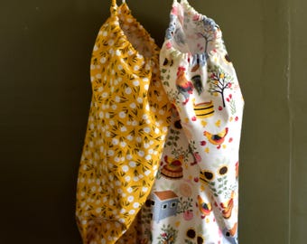 May include: Two fabric produce bags hang from a wooden rack. One bag is yellow with a white floral pattern. The other bag is white with a colorful farm scene featuring chickens, trees, and a small house.