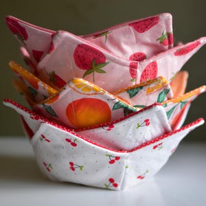 May include: A stack of four fabric bowl cozies with fruit patterns. The top cozy features strawberries on a pink background. The next has a peach design, followed by a cherry pattern. Each has a contrasting red trim.