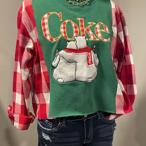 May include: A green sweatshirt with red and white plaid sleeves and a raw hem. The front features the word Coke in red and white checkered lettering above an embroidered graphic of two white polar bears wearing a red scarf and holding bottles.