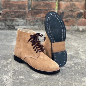 Könnte beinhalten: Hellbraune Wildleder-Ankle Boots mit dunkelbraunen Schnürsenkeln und schwarzer Sohle. Ein Stiefel steht aufrecht, der andere wird mit der Sohle nach oben gezeigt, wodurch die Nähte sichtbar werden. Die Stiefel stehen vor einer Backsteinmauer.