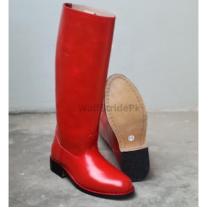 May include: A pair of bright red knee-high boots with a smooth finish, featuring a low black heel and a tan leather sole. The text WolfStridePk is visible on the side of the upright boot, and a small white size sticker is on the sole.