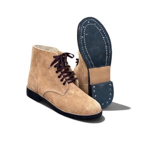 May include: A pair of tan suede ankle boots with dark brown laces. One boot is shown from the side, while the other is positioned behind it, displaying the dark blue rubber sole with visible stitching and a stacked heel construction.