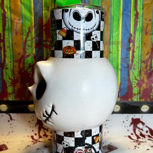 May include: A unique tumbler featuring a Jack Skellington design. The top section has a black and white checkerboard pattern with Jack's face, pumpkins, and candy corn. The bottom is a white skull shape. A green apple-shaped lid and a straw complete the design.