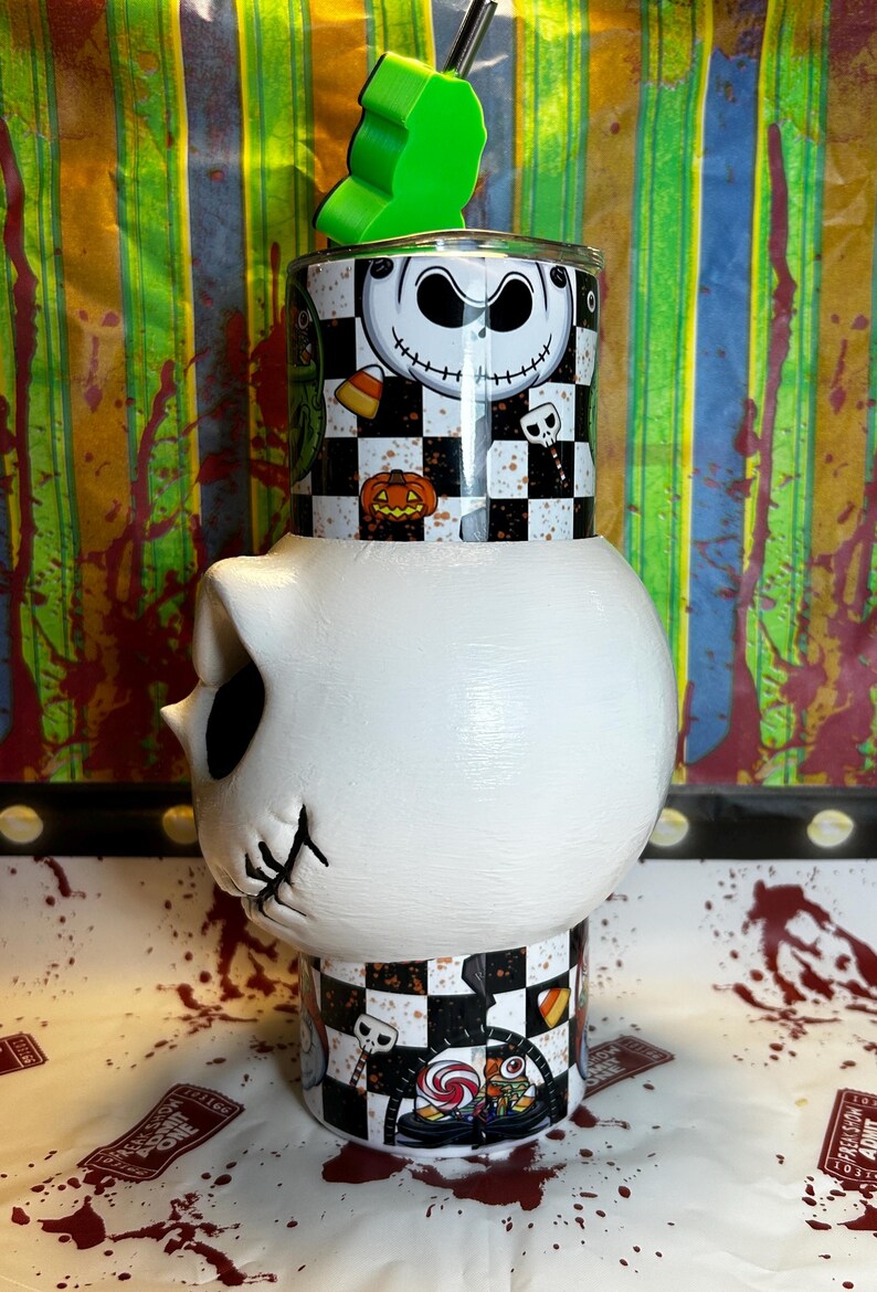 May include: A unique tumbler featuring a Jack Skellington design. The top section has a black and white checkerboard pattern with Jack's face, pumpkins, and candy corn. The bottom is a white skull shape. A green apple-shaped lid and a straw complete the design.