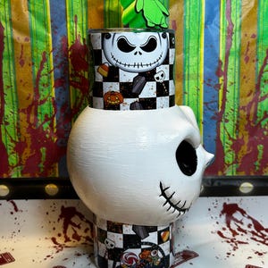 May include: A white, Jack Skellington-themed tumbler with a green Oogie Boogie topper. The tumbler features a black and white checkered pattern with Halloween-themed graphics. The design includes a Jack Skellington face, pumpkins, and tombstones.