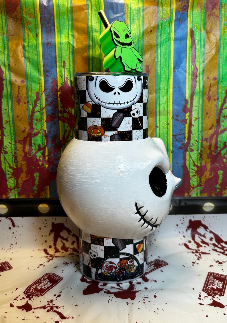 May include: A white, Jack Skellington-themed tumbler with a green Oogie Boogie topper. The tumbler features a black and white checkered pattern with Halloween-themed graphics. The design includes a Jack Skellington face, pumpkins, and tombstones.