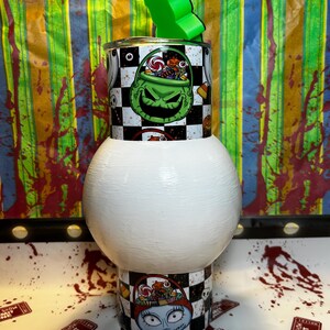 May include: A Halloween-themed tumbler with a green and black design featuring a grinning character and a checkerboard pattern. The tumbler has a white base and a green straw holder. The design includes images of candy and a character from the movie.