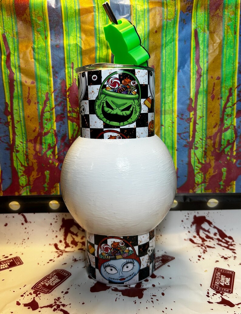 May include: A Halloween-themed tumbler with a green and black design featuring a grinning character and a checkerboard pattern. The tumbler has a white base and a green straw holder. The design includes images of candy and a character from the movie.