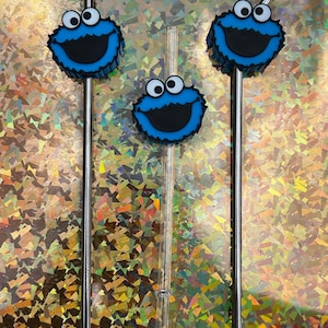 May include: Three drinking straws, two stainless steel and one clear plastic, with Cookie Monster character toppers. The toppers are blue with white eyes and a black mouth. The straws are for drinks.