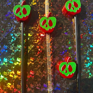 Op de afbeelding: Vier herbruikbare rietjes met appelvormige bedels. De bedels zijn rood en groen met een schedelachtig ontwerp. Twee rietjes zijn zwart en twee zijn transparant. De achtergrond is een glanzend, iriserend oppervlak.
