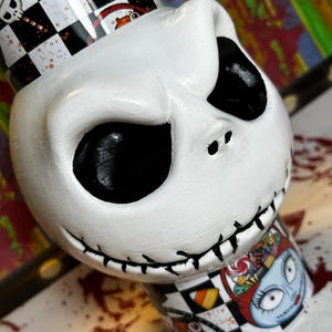 May include: A white, hand-painted Jack Skellington figurine from The Nightmare Before Christmas. The figurine features a stitched mouth, black eye sockets, and a checkered pattern with other characters. The base has a Sally illustration.