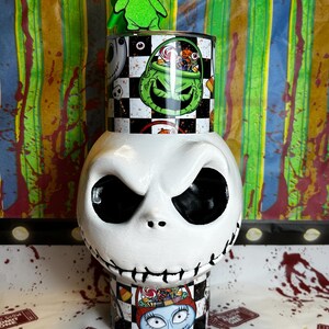 May include: A white ceramic Jack Skellington head with black eyes and stitched mouth. A cylindrical container with a checkered pattern and Oogie Boogie and Sally designs sits on top. A green Oogie Boogie figure is on top.