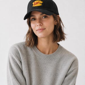 May include: A black baseball cap featuring an embroidered yellow oval patch with the red text Luke's. The cap has a curved brim and a structured crown, constructed from a solid black fabric with matching stitching throughout the design.