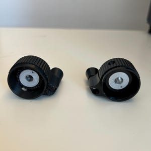 Può includere: Due parti meccaniche nere e circolari con un bordo esterno scanalato e un centro color argento. Ogni parte ha un piccolo braccio curvo che si estende dal lato. Le parti sono poste su uno sfondo chiaro e uniforme.