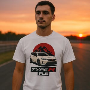 Puede incluir: Camiseta blanca con un gráfico de un coche blanco, un diseño de montaña y sol rojos. El texto "TYPE R FLS" está debajo del coche. La camiseta se lleva contra un atardecer naranja.