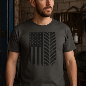 May include: Dark gray t-shirt featuring a distressed American flag design combined with tire tread marks. The graphic is in black. The shirt is a crew neck and short-sleeved.