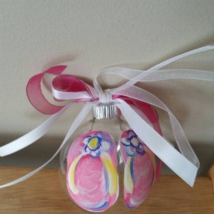 May include: A glass ornament with a hand-painted design of pink flip flops with yellow straps and blue and purple flowers. The ornament is tied with a white ribbon and a pink ribbon.