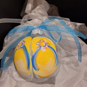 May include: A clear glass Christmas ornament with a hand-painted design of yellow flip flops with blue straps and white flowers. The ornament is tied with a blue and white ribbon.