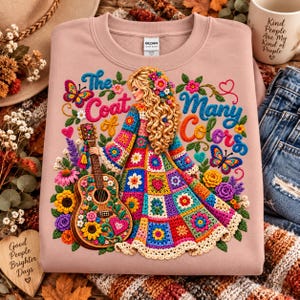 Peut inclure: Un sweat-shirt rose clair avec une illustration d'une personne aux longs cheveux blonds bouclés portant un manteau style crochet coloré. Le texte indique The Coat of Many Colors. Une guitare ornée de fleurs et d'un cœur accompagne la figure.