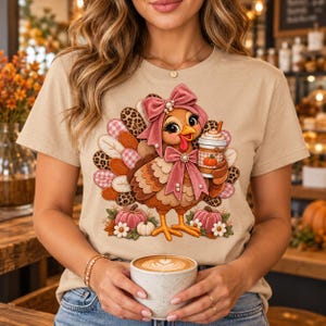 Peut inclure: Un t-shirt beige avec un motif de dinde de dessin animé portant un nœud rose et tenant une tasse de café. La dinde a des plumes aux motifs léopard et écossais rose. Des citrouilles roses et des fleurs blanches sont à la base.