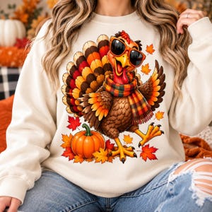 Peut inclure: Un sweat-shirt couleur crème arborant un motif de dinde portant des lunettes de soleil noires et une écharpe à carreaux. La dinde se tient à côté d'une citrouille orange entourée de feuilles d'érable rouges, orange et jaunes.