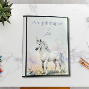 Könnte beinhalten: Eine weiße Zeugnismappe mit einem aufgedruckten Bild eines weißen Einhorns auf einer Blumenwiese. Der Schriftzug Zeugnismappe und der Name Lea sind in dunkelgrauer Schrift im oberen Bereich des Einbands zu sehen.