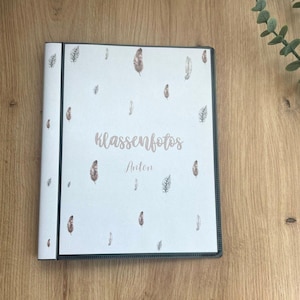 Könnte beinhalten: Ein weißes Fotoalbum mit schwarzem Rücken und Rand, verziert mit einem Federmuster. Auf dem Cover steht der Text "Klassenfotos" und "Anton" in einer zarten Schrift. Das Album liegt auf einer Holzoberfläche.
