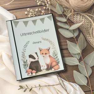 Könnte beinhalten: Ein weißes Fotoalbum mit dem Text "Ultraschallbilder" und dem Namen "Amira". Das Cover zeigt Illustrationen eines Dachses, eines Fuchses und Pilzen. Oben befindet sich ein Banner mit grauen Flaggen.