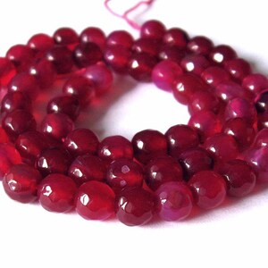 Può includere: Un filo di perline sfaccettate di pietre preziose rosso intenso. Le perline sono rotonde e hanno una superficie lucida e riflettente. Il filo è di colore rosa chiaro. Le perline sono disposte in un motivo circolare su sfondo bianco.