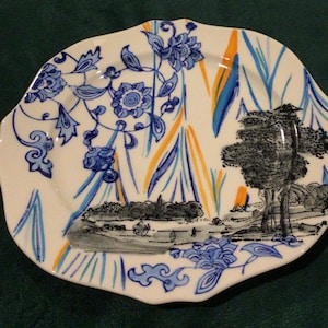 May include: A white ceramic plate with a scalloped edge features a blue and white floral pattern, a black and white landscape scene, and yellow and orange accents.