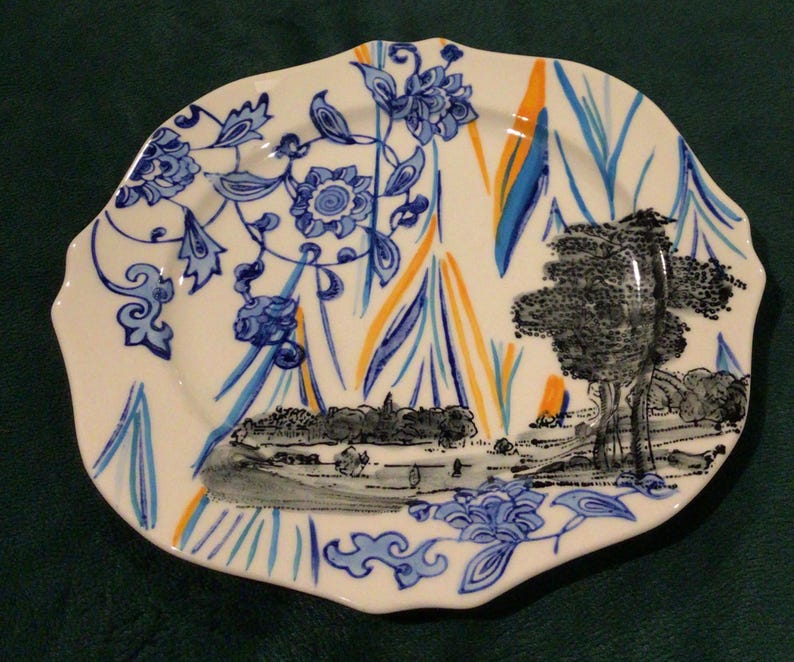 May include: A white ceramic plate with a scalloped edge features a blue and white floral pattern, a black and white landscape scene, and yellow and orange accents.