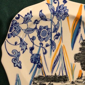 May include: A white ceramic plate with a blue and yellow floral design. The plate has a scalloped edge and a black and white image of a landscape at the bottom.