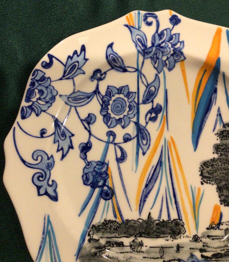 May include: A white ceramic plate with a blue and yellow floral design. The plate has a scalloped edge and a black and white image of a landscape at the bottom.