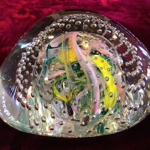May include: A clear glass paperweight with an internal design of swirling yellow, pink, and green colors. The paperweight is oval-shaped and filled with numerous small air bubbles, creating a decorative effect.