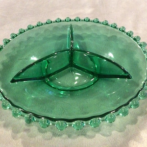 May include: A vintage green glass serving dish with three compartments. The oval-shaped dish has a scalloped edge with small, round, raised details. The interior is divided into sections, perfect for serving snacks or appetizers.