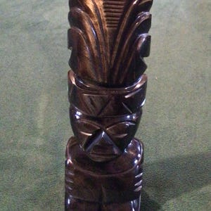 May include: A black obsidian tiki statue with intricate carvings. The statue is standing on a green surface.