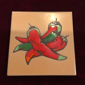 May include: Square ceramic tile featuring a vibrant design of red and green chili peppers. The peppers are arranged in a cluster against a tan background, with a dark outline. A decorative item for the kitchen or dining area.