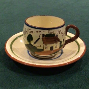 May include: A ceramic teacup and saucer set. The cup features a hand-painted scene of a house and tree with the text "Walton-on-the Naze". The saucer has a green and brown rim. The cup has a brown handle.