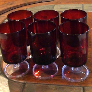 May include: A set of six deep red glass goblets with clear glass stems and circular bases. Each goblet features a cylindrical bowl with a smooth, uniform surface and a consistent dark ruby color throughout the glass structure.