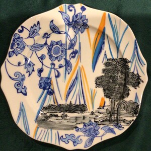 May include: A white ceramic plate with a blue and orange floral design. The plate has a scalloped edge and a black and white landscape scene in the center.