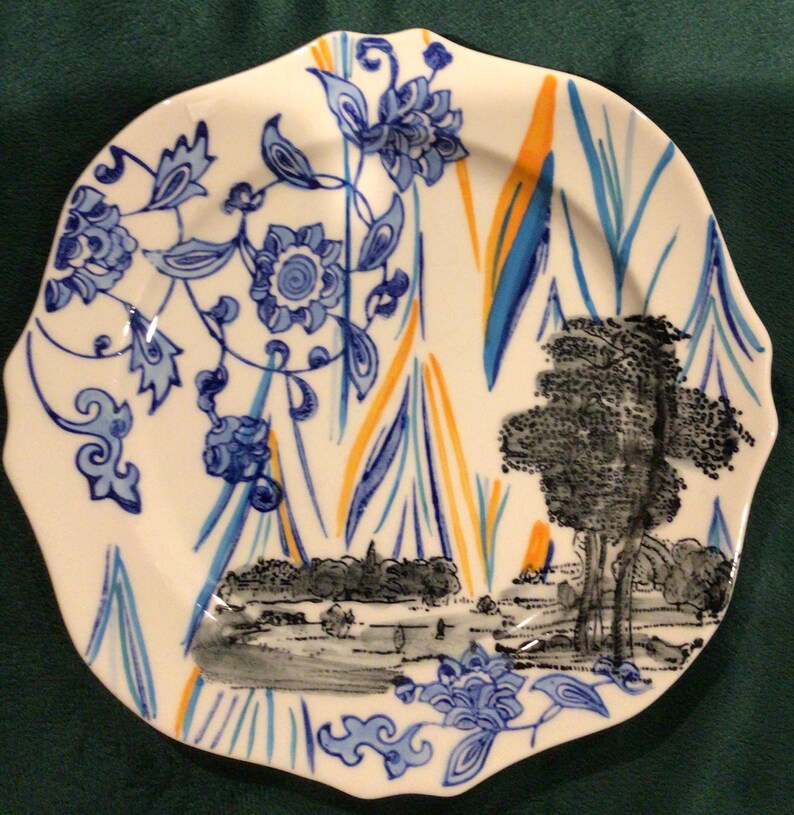 May include: A white ceramic plate with a blue and orange floral design. The plate has a scalloped edge and a black and white landscape scene in the center.