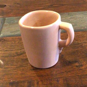 May include: A solid light pink ceramic mug with a smooth, glossy finish. The mug features a cylindrical body with a slightly flared rim and a sturdy, curved handle attached to the side. The interior and exterior surfaces are a uniform pastel pink.