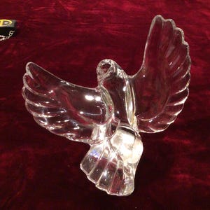 May include: A clear glass dove sculpture with outstretched wings. The bird is detailed with etched feather patterns and sits on a dark red surface. The sculpture is a decorative item.