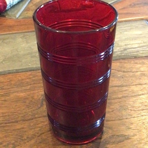 May include: A cylindrical drinking glass made of deep red translucent glass. The exterior surface features a series of horizontal, raised circular bands or rings that wrap around the body of the glass, creating a textured, ribbed appearance.